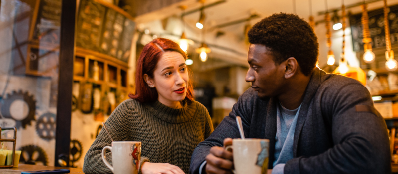 A young woman and man sit at a coffee shop. The woman has toxic behaviors that affect their relationship and her partner.