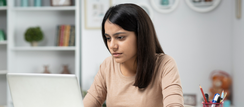 A high school female in a beige shirt on her computer. She is managing college application stress. 