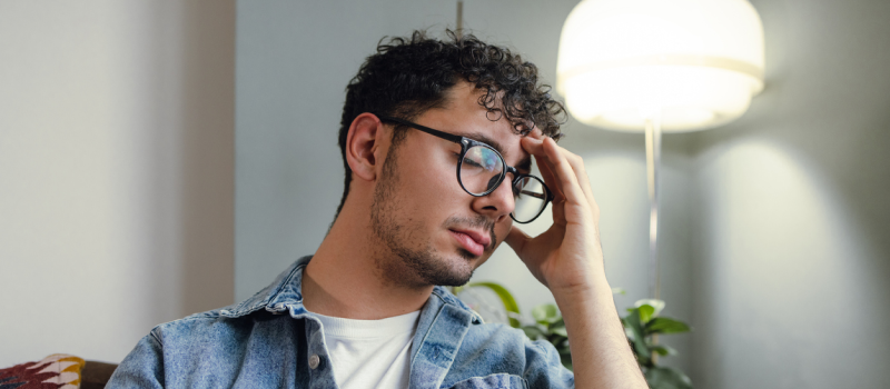A young male sits in his living room wearing a jean jacket and white shirt. He is experiencing an emotional hangover from a traumatic or otherwise draining emotional encounter.