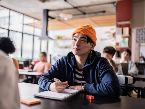 A high school student at their desk in class in a blue jacket and orange hat. He is managing college application stress.