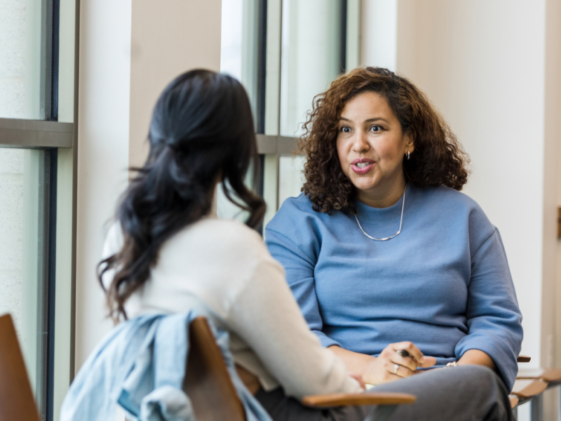 A therapist sits with a young woman. The therapist is in a blue shirt, and is addressing mental healthcare barriers for Hispanic/Latinx clients inside and outside the therapy room.