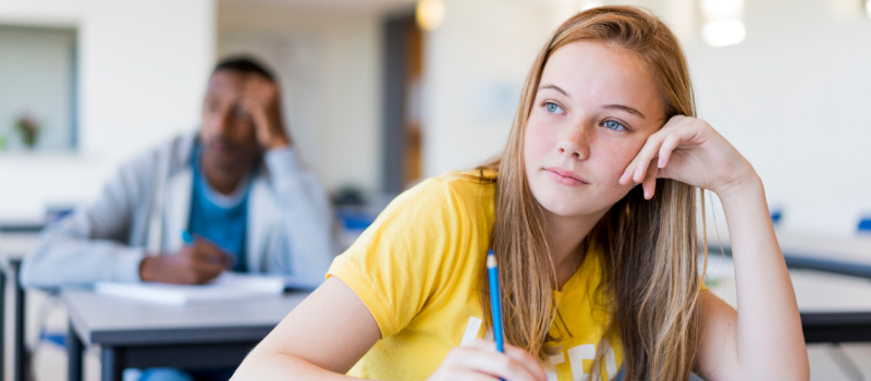 A high school student sits at her desk. She has on a yellow shirt and is holding a pencil.