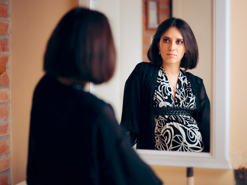 A pregnant person wearing a black and white dress and a black sweater looks in a mirror in the bathroom and wonders if her narcissistic personality disorder will be genetically passed to her child.