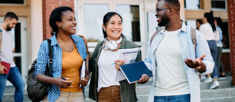 A trio of college students who are prioritizing their mental health stand in front of a campus building holding backpacks and books and smiling.