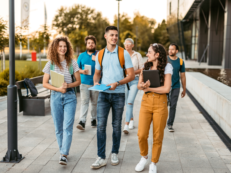 A group of college students carrying books and backpacks walk down a campus road smiling. They are prioritizing their mental health and enjoying their time as college students.