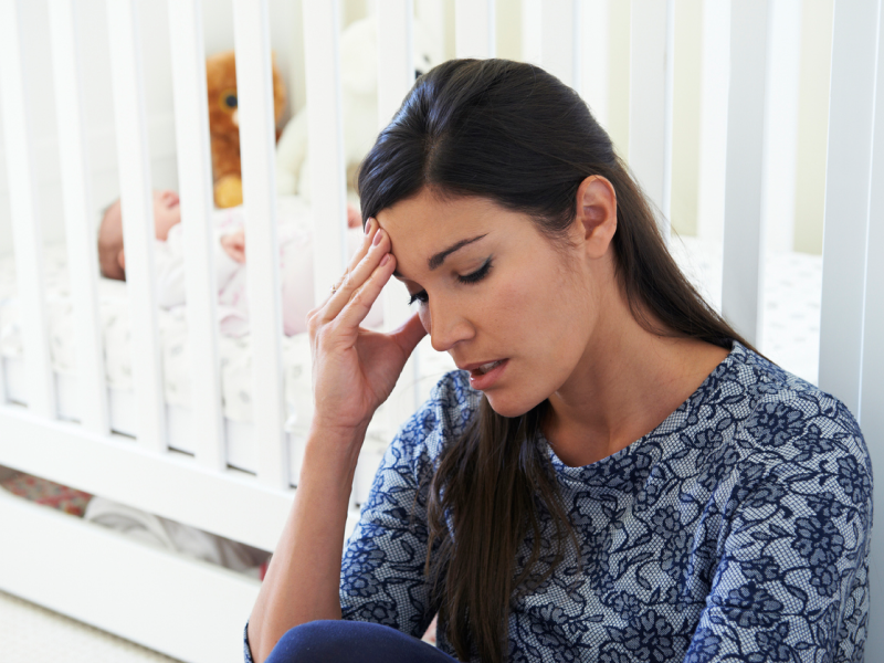 A new mom sits next to the crib of her recently born sleeping baby. She puts her hand on her forehand and closes her eyes in a moment of overwhelming worries about the baby's sleeping and eating. She is dealing with new mom anxiety.