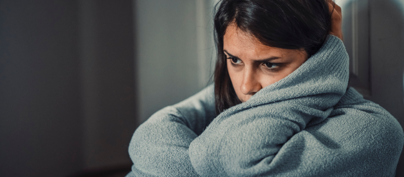 A woman dealing with postpartum OCD sits in a dark room in a sweater holding her head and frowning as she contends with the postpartum OCD symptoms.