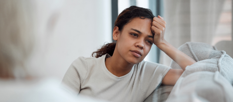 A woman in a white shirt sits on a therapist's couch discussing somatic flashbacks she's been experiencing recently due to past trauma.