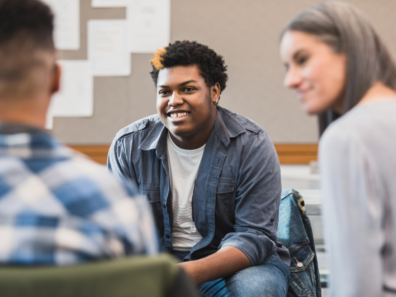 A group of college students sit in a classroom talking. They are managing their mental health well with the support of mental health programs, including Charlie Health.