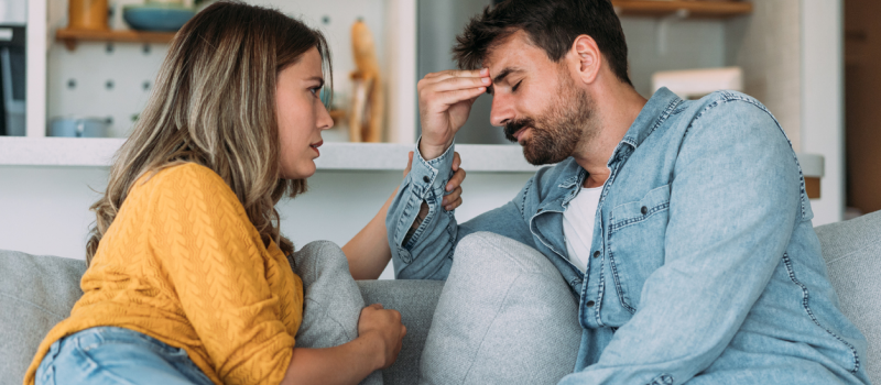 A woman in a yellow shirt and a man in a denim shirt sit on a couch during an argument. The man grasps his head trying to make sense of what the woman, who has narcissistic personality disorder, is saying.