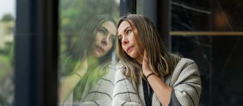 A woman dealing with comorbid PTSD and anxiety puts her hand on her neck and leans against a window looking out at the forest to soothe herself from trauma and anxiety symptoms.