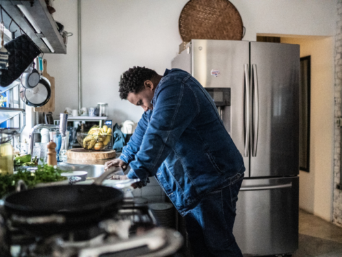 A man wearing a blue long sleeve shirt stands over the sink in his kitchen feeling overwhelmed as he deals with post-traumatic stress disorder symptoms.