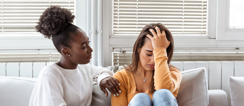 A young woman in a white sweater sits with her friend in an orange top. The woman is comforting her friend, who has experienced chronic trauma in her life.