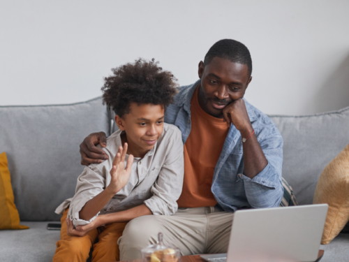 A father and son sit on their couch on the computer. The son is in Charlie Health's virtual IOP, which new research shows reduces mental health-related ED admissions.