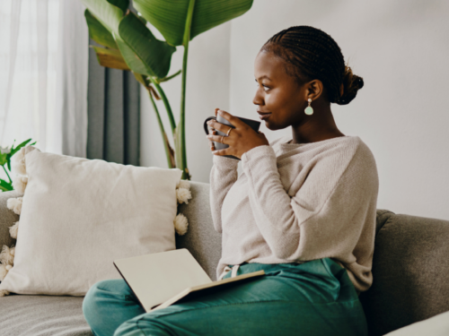 A young woman sits on her couch drinking coffee. She is doing CBT journaling to help deal with negative thoughts.