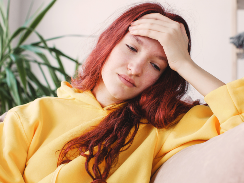 A young woman in a yellow sweatshirt sits at home. She is having ADHD burnout and has been tired all the time.