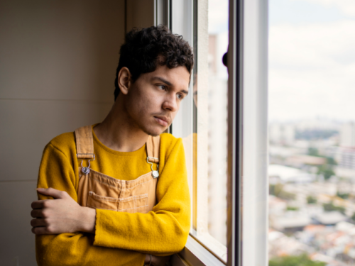 A young man in an orange top and overalls looks outside of his window. He has experienced different types of trauma in his life.