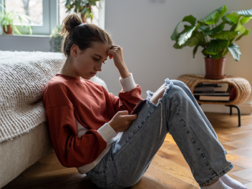 A female teenager sits on the floor of her room. She is looking at an emotional wheel on her phone to understand her emotions.