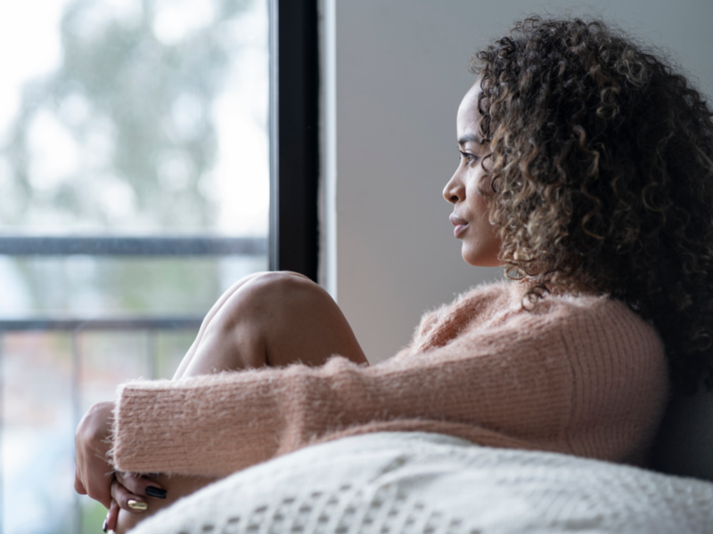 A young woman has PTSD from childhood abuse as an adult. She sits at home staring out the window.