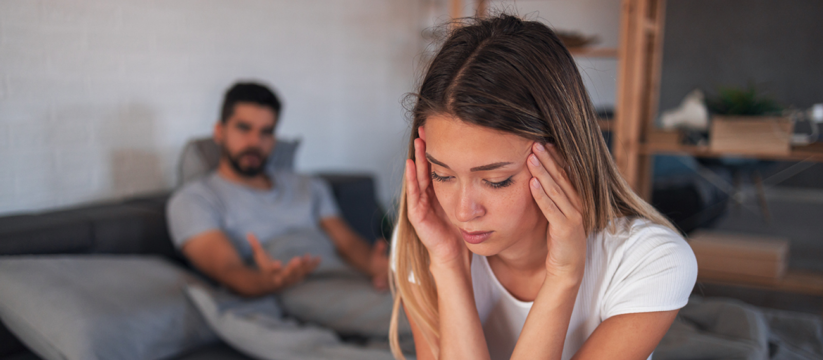 A young couple sit on their couch at home. The male gaslights his partner, and she is experiencing increased guardedness and increased mistrust of others.