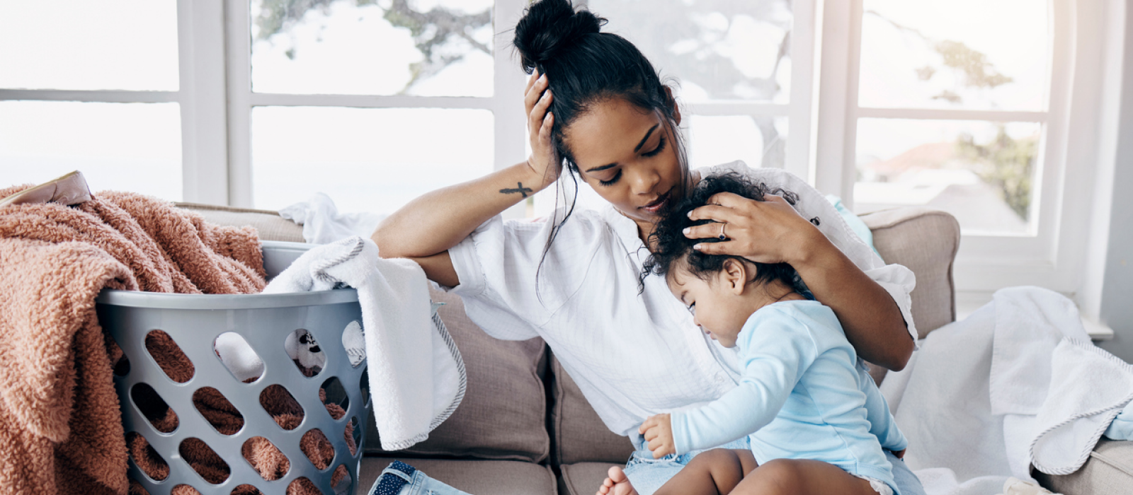 A young mom sits on her couch with her child and a lot of laundry. She is experiencing mom burnout and is considering additional approaches to parenting.