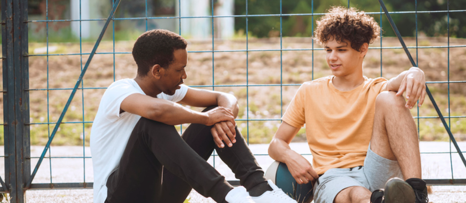 Two young men sitting on the ground at the basketball court. One of them is coping with a depressive episode and is using their support system to help them.
