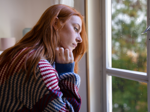 A young woman looks out of her window at the changing seasons. She uses 7 therapist-approved tips for seasonal depression.
