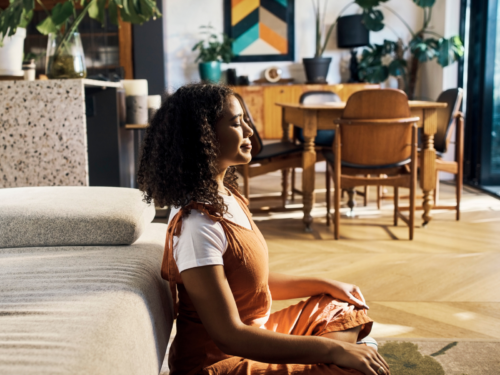 A young woman sits at home on her floor. She is practicing somatic breathwork to help her chill out.