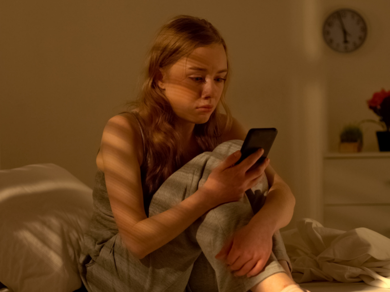 A young girl sits in bed on her phone. She is experiencing depression following a breakup.