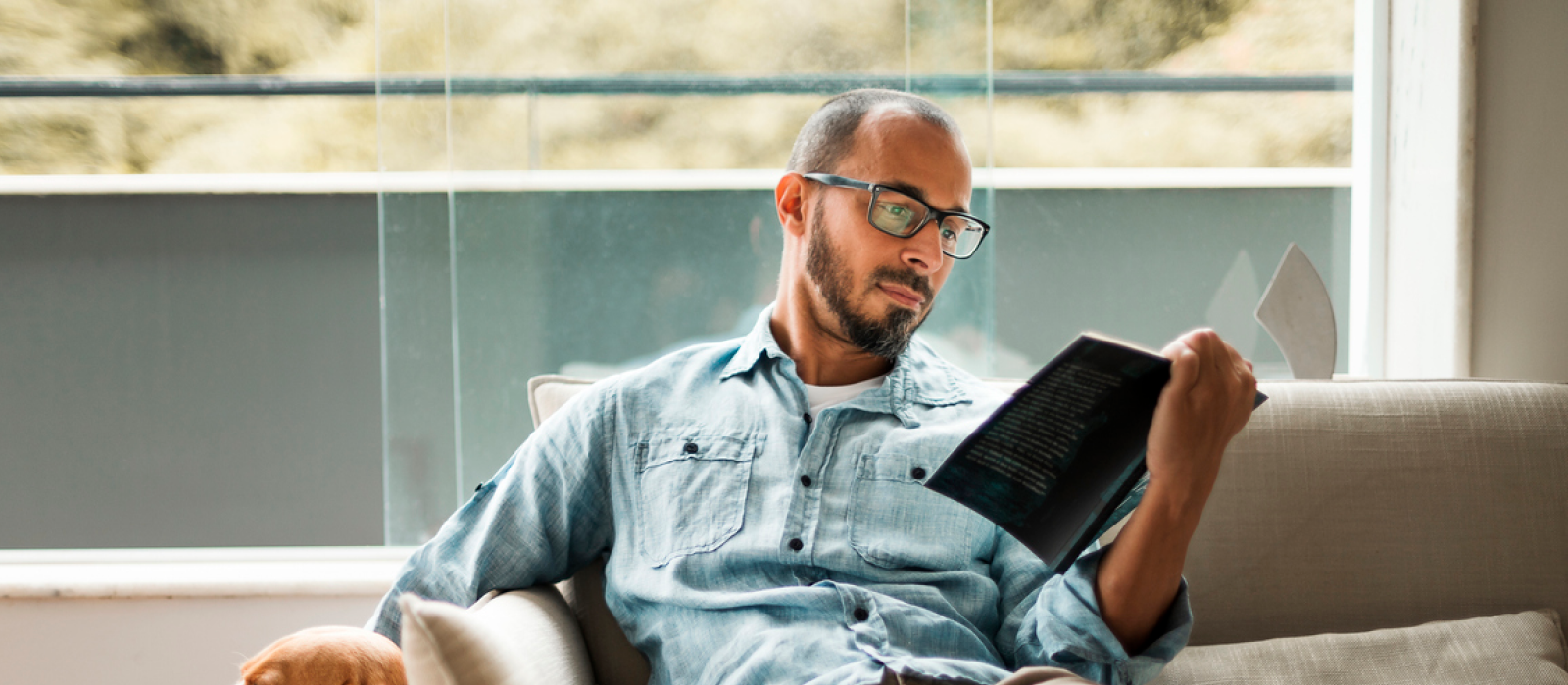 A young man is practicing self-care by reading on his couch to reduce or prevent compassion fatigue and burnout.