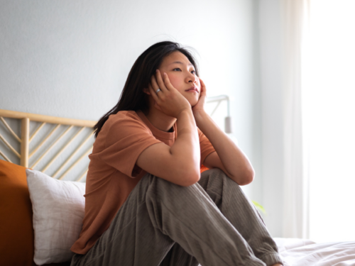 A young woman sits on her bed. She feels herself in survival mode, which is leading to chronic stress and exhaustion.