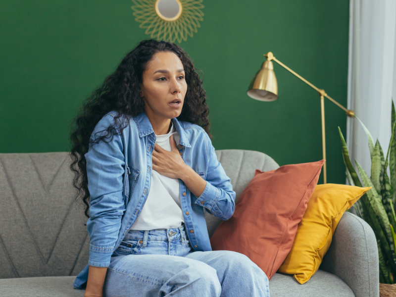 A person sits on a couch and puts her hand on her chest to calm shortness of breath during an anxiety spell.