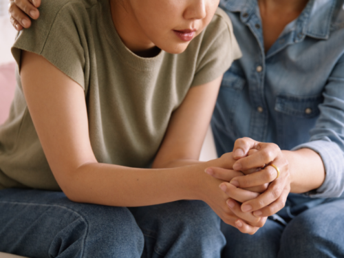 Two people sit on a couch. One has been self-harming and the other is holding her hands discussing how DBT for self-harm could help her manage symptoms.