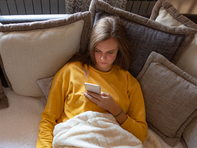 A young girl in a yellow sweatshirt, a picture of the youth mental health crisis, looks at social media on her phone.