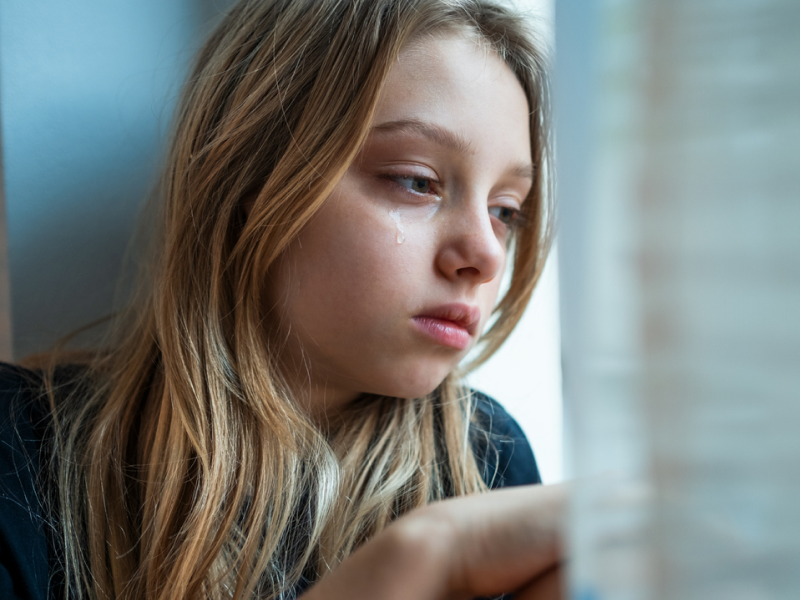 A young girl looks out a window in grief. She could use DBT to cope with grief.