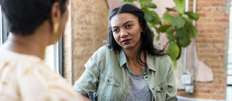 A woman with black hair and a green and grey shirts sits in the background as her therapist sits in the foreground offering therapy for self-esteem.