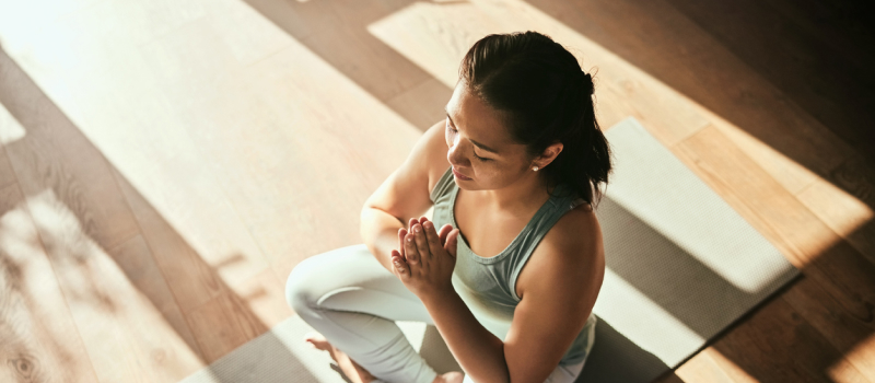 A woman sits on a yoga mat with legs crossed and arms in front of her practicing trauma-informed yoga.