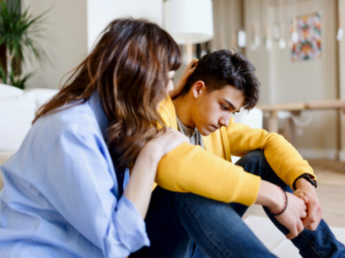 A mother in a blue shirt comforts her son who is sitting on the ground in a yellow shirt dealing with the effects of reactive attachment disorder.