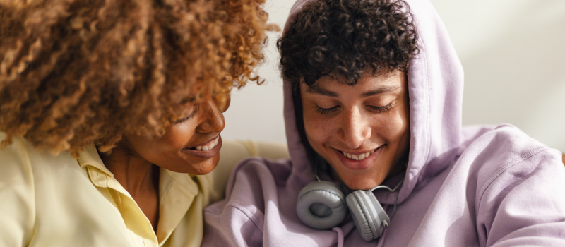 A young person who has been in therapy and processed the effects of growing up with a bipolar parent sits on a couch smiling next to a friend.