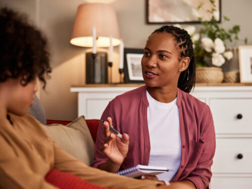 A clinician in a red sweater sits in a therapist's office talking with a client about the results of her C-SSRS questionnaire.