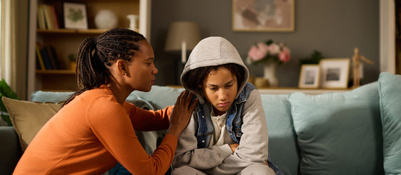 A mom in an orange shirt puts her arms around a teen in a grey sweatshirt on a teal couch who is struggling with reactive attachment disorder.