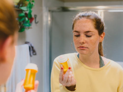 A young woman wearing a yellow shirt holds a bottle of medication considering whether to begin SSRIs or SNRIs.