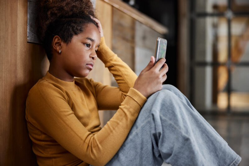 Sad child sitting on floor looking at phone, illustrating childhood sadness and the importance of parental support.