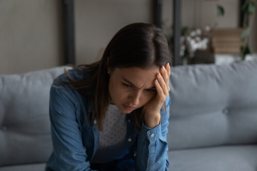 Exhausted frustrated young woman sit on couch at home hugging head due to pain panic attack feeling overwhelmed with her drinking problem.