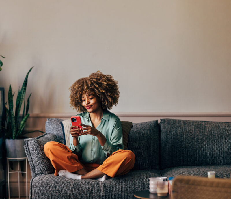 A smiling elegant African-American female using her smartphone to create her DBT while sitting on the cozy sofa in the living room.