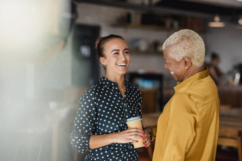 Two women colleagues laughing while standing in a cafe at their workplace. They feel protected from harassment in the workplace.