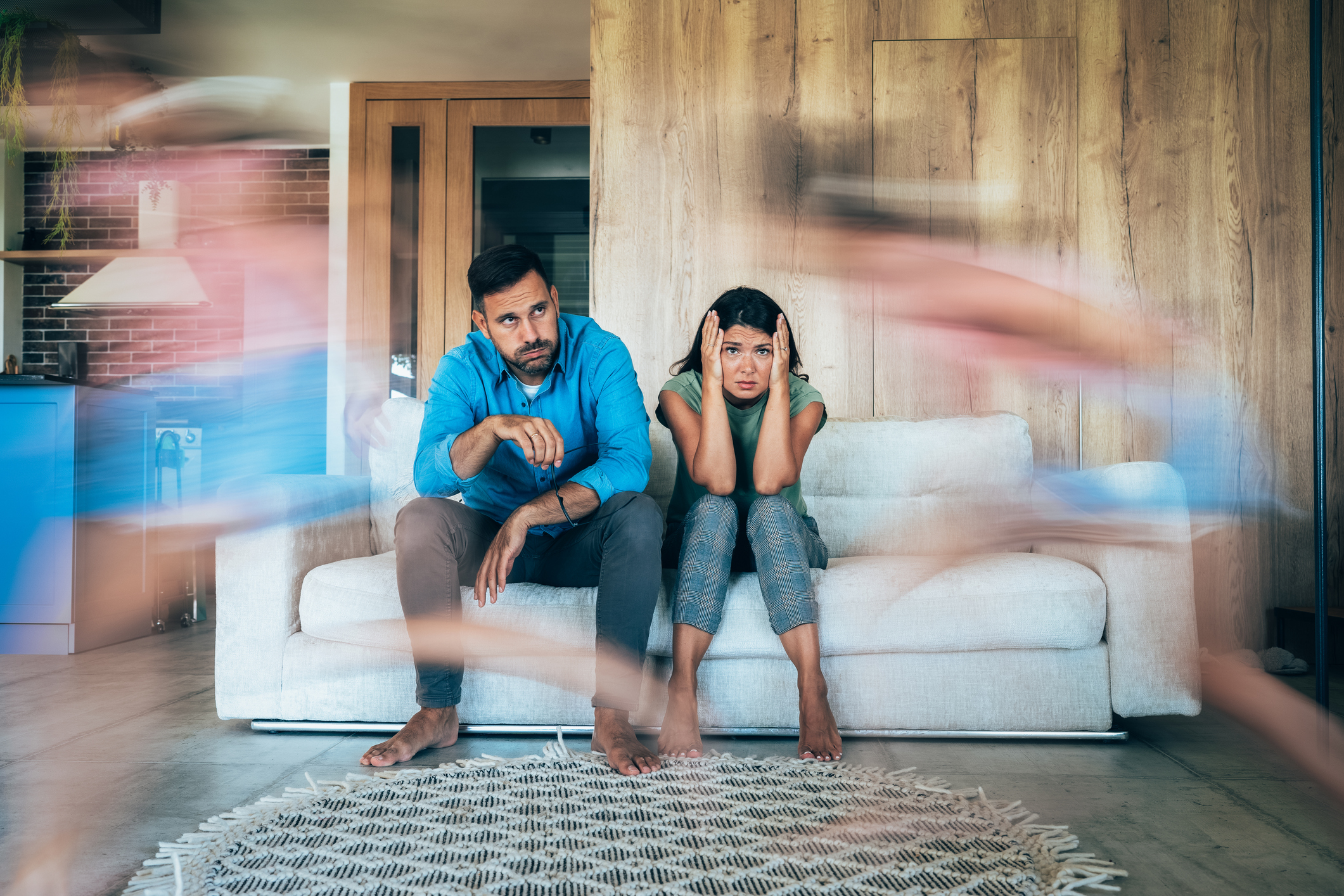 Tired mother and father sitting on couch feels annoyed and exhausted while child with conduct disorder runs around the sofa.