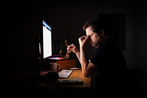 Tired and stressed man illuminated by the glow of their computer screen in a dark room at night while working late. Burn out.