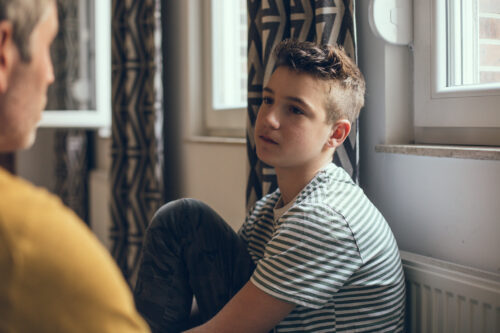 Teen boy sitting with knees pulled up, listening attentively during a serious conversation, possibly in a therapy or support setting.