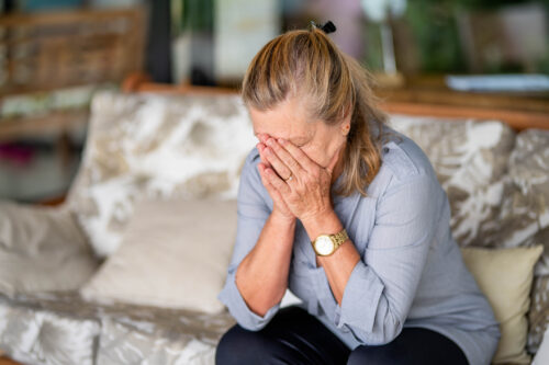 Older woman sitting on a couch with her face in her hands, appearing distressed or overwhelmed, possibly experiencing emotional pain or trauma.